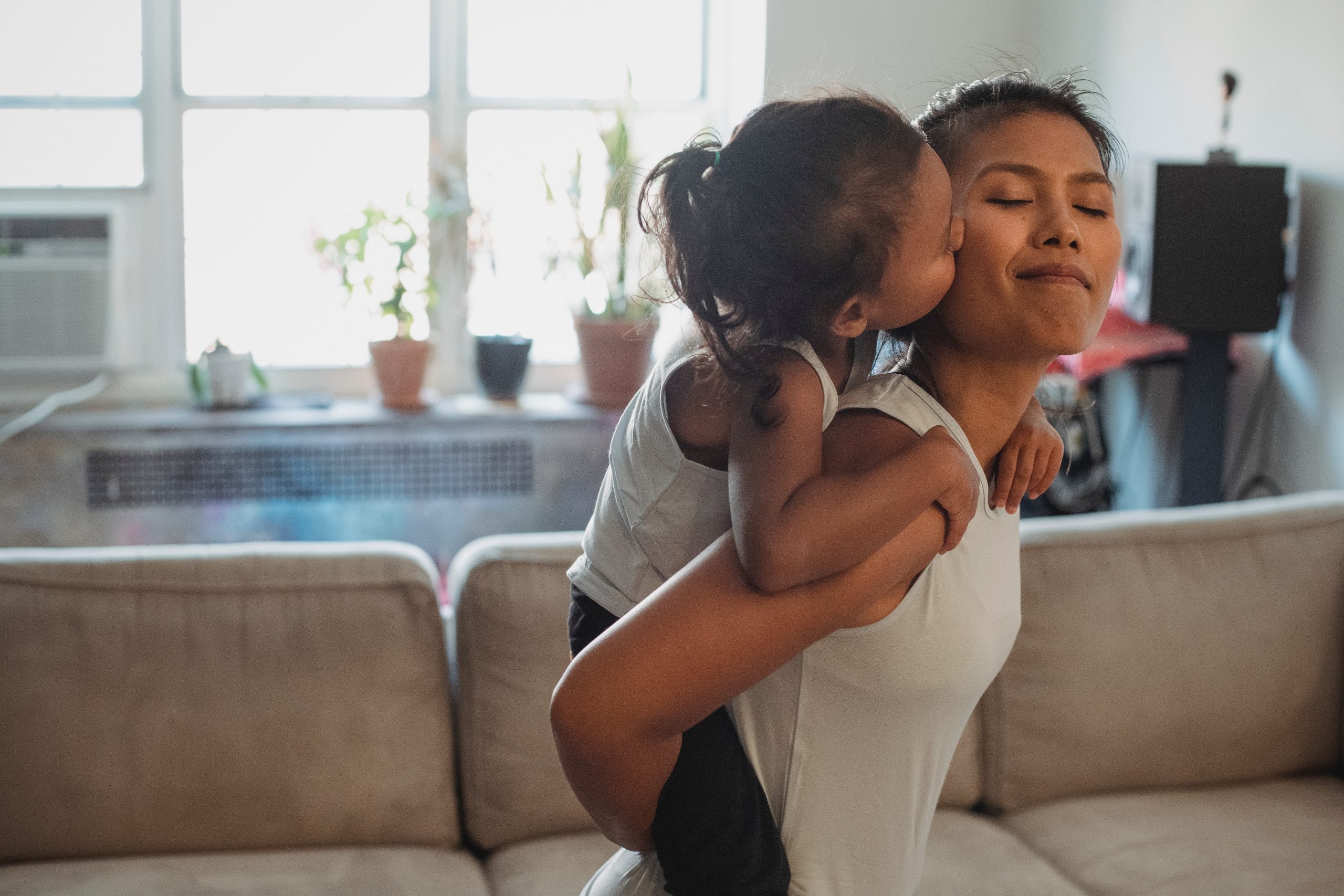 Photo of a young parent with their eyes closed, smiling, and holding their preschool age child. The child is kissing their parent